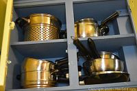 Cabinet containing various metal cookware items including perforated pot, saucepans, and frying pans with black handles