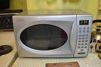 Front angled view of silver Sunbeam microwave oven on countertop next to stove. Shows keypad with numeric and function buttons and digital display.