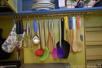 Assorted kitchen utensils hanging on rod including spatulas, ladle, whisk, masher, slotted spoon in colorful and metal finishes