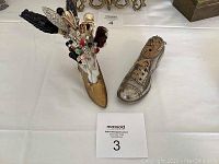 Two metal shoe-shaped pincushions: a gold ladies' shoe and a silver men's brogue shoe, both holding many hat pins.