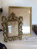 Two antique brass picture frames shown upright on a table, one large etched rectangular frame and one smaller ornate frame with scroll and floral decoration.