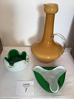 Angled view of amber glass pitcher next to two green and white cased glass ashtrays