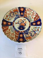 Top-down view of scalloped-edge porcelain dish showing bold Imari-style floral and geometric motifs in cobalt blue, iron red, and gold highlights.