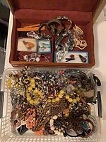 Open wooden box with various bracelets and brooches inside, plus plastic basket with colorful vintage necklaces and other costume jewelry piled inside it.