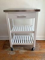 Front view of the white metal kitchen trolley with stainless steel top, drawer under top, two slatted shelves, and black locking caster wheels.
