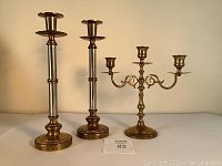 Photo showing one pair of tall brass candlesticks and a triple-arm brass candlestick on a neutral surface against a beige wall.