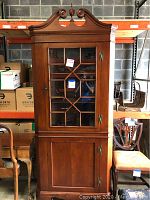 Full view of a wooden corner hutch with a glass front door and a wooden door below, both with antique style handles and keyholes.