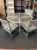 Full set showing round white metal patio table with glass top and four blue metal frame chairs with white woven straps