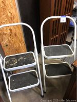 Two white metal folding 2-step stepstools with black non-slip surfaces on each step, positioned side by side showing front and top views.