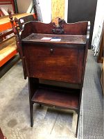 Wooden secretary desk closed showing front panel, bottom shelf, and decorative medallion on top with some damage.
