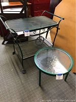 Green metal two tier tea cart with mesh shelves and wrought metal handles, alongside green metal round end table with glass top