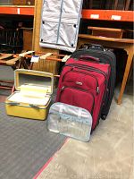 A group of luggage cases including red and black rolling fabric suitcases, a yellow hard toiletry/train case, and a small silver case in front of the luggage racks.