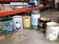 Seven assorted trash cans with various decorative designs in metal and one medium wooden bin.