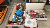 Plastic bin filled with holiday wrapping paper rolls, gift bows, and ribbon beside a box with silver snowflake ornaments.