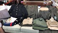 Photo of assorted military clothing including dark navy jacket with gold buttons, several coats and jackets in olive green and khaki, various shirts, and folded pants on a couch.
