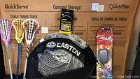 Photo shows boxed Quick Play table tennis table with paddles and partially visible lacrosse sticks in front, Easton net partially visible, snowboard propped upright to the right.