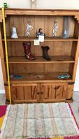 Front view of wooden bookcase with measuring tape stretched showing approximate height 5 feet, three upper shelves holding Southwestern decorative items including kokopelli figurines, ceramic bell, cowboy boots, and two turquoise lizards on bottom open shelf.