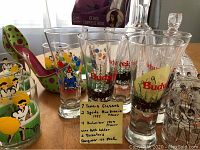Photo showing various glasses including tennis-themed highball glasses, Spudz Mackenzie pilsner glasses, Budweiser pilsner glasses, high heel wine bottle holder and decanters highlighting lot contents.