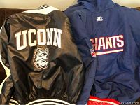 Photo showing back of two jackets: black and white UConn Huskies leather varsity jacket and blue New York Giants jacket with NFL logo.