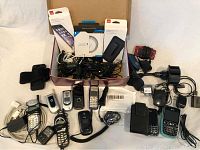 A box with multiple cell phones including flip phones, Blackberry smartphones, feature phones, with accompanying chargers, holsters, and cables.