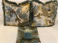 Two decorative square pillows with floral pattern and green fringe trim placed with a green-toned glass bowl featuring textured radiating lines.
