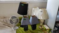 Five table lamps and one desk fan displayed on a green surface near a window.