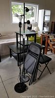Two black floor lamps with adjustable lamp heads and cords at base. Four black metal folding chairs with upholstered seats and backs leaning against each other near the lamps. Concrete floor, daylight through window.