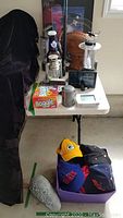 White folding table with assorted hats in purple bin, Boggle game, stainless steel container, digital devices, two metal lanterns, carved wood football, and framed Patriots poster in background