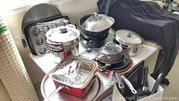 Multiple pots and pans including a steamer with lid, roasting pans, frypans, and bakeware on a table