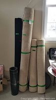 Several upright rolled carpets and flooring remnant in corner near window
