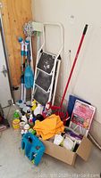 Photo showing three-step folding stepladder leaning against a wall surrounded by various cleaning tools and supplies including mops, brushes, bucket, and cleaning liquids.