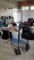 Photo showing the white metal clothes drying rack and black metal shopping cart from a side angle in a garage setting with various items around.