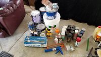 Photo showing an assortment of paint cans of different sizes, caulking tubes, two caulking guns, and small maintenance bottles arranged on the floor.