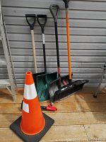 View of four snow shovels with different handle colors and blade types, plus an orange safety cone with reflective strip and black base.