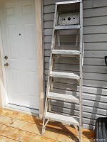 Full view of the six step aluminum ladder standing upright outside near a door, showing some wear and marks on the surface.