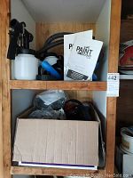 Photo of wooden shelving unit showing the overall lot contents with paint sprayer, paint containers, manuals, and safety equipment in a box.