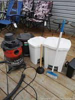 Overall shot showing the Shop-Vac vacuum, three white plastic step-on waste cans, broom, and plunger on a wooden deck.