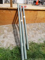 Three green adjustable metal jack posts leaning against a wooden deck outside on gravel and grass surface.