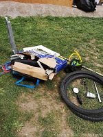 Photo showing discarded items on grass including folded cardboard pieces, bike tires, and scattered bicycle parts (mirror, light, kickstand)