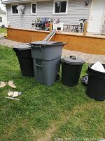 Wide image showing all four garbage cans including one with wheels, set on grass against house background.