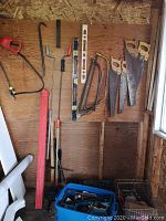 Wide view of tools mounted on wooden wall including hand saws, crowbar, levels, and plumber's snake. Containers of fittings on floor.