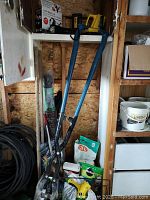 Post hole digger with blue handles and black blades, garden supplies including Miracle Gro boxes and additional gardening tools.