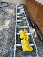 Full length of the aluminum extension ladder resting on gravel with the yellow Roofers Worlx ladder mount attached on a rung near the base.