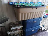 Stacked Rubbermaid bins with wrapping paper on top and two of the Christmas-themed cardboard boxes beside them.