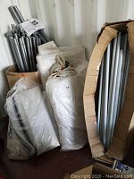 Grey powder-coated steel frame poles and connectors standing upright next to bundled canopy components in plastic bags