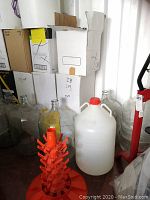 Wide shot of 5 glass carboys clustered against wall with 1 large white plastic jug and orange bottle dryer in front