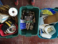 Top-down view of three green plastic bins containing various household hardware and tools including electrical cords, gloves, plumbing fittings, and tape measure.