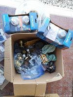 Box with various door hardware: two Schlage lock sets in blue packaging, assorted keys, hinges, door closer arm, and additional metal hardware parts visible.