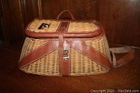 Side view of vintage wicker fishing creel showing woven wicker body and leather straps with buckle closure.