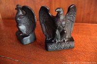 Front and side view of two cast metal American eagle bookends with detailed feathers and vine patterned bases on a wood surface.